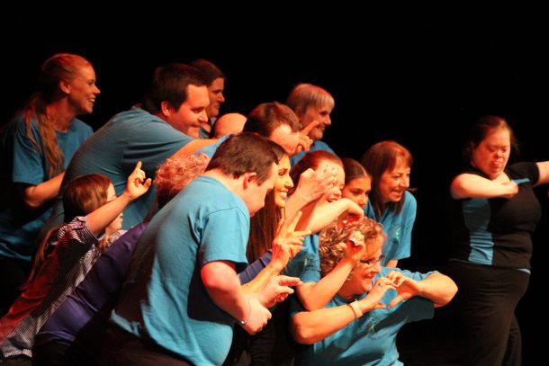 A group of smiling people in blue T-shirts, the All Abilities Dance Troupe, gathers together and makes heart shapes with their hands.