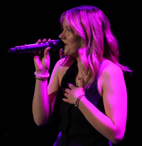 A young woman with blonde hair in purple light sings into a microphone