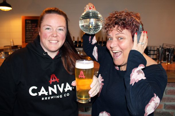 A woman in a black hoodie that says Canvas Brewing Co. holds a glass of beer, and a woman in a black and pink shirt dances and holds a small sparkling disco ball while wearing headphones.