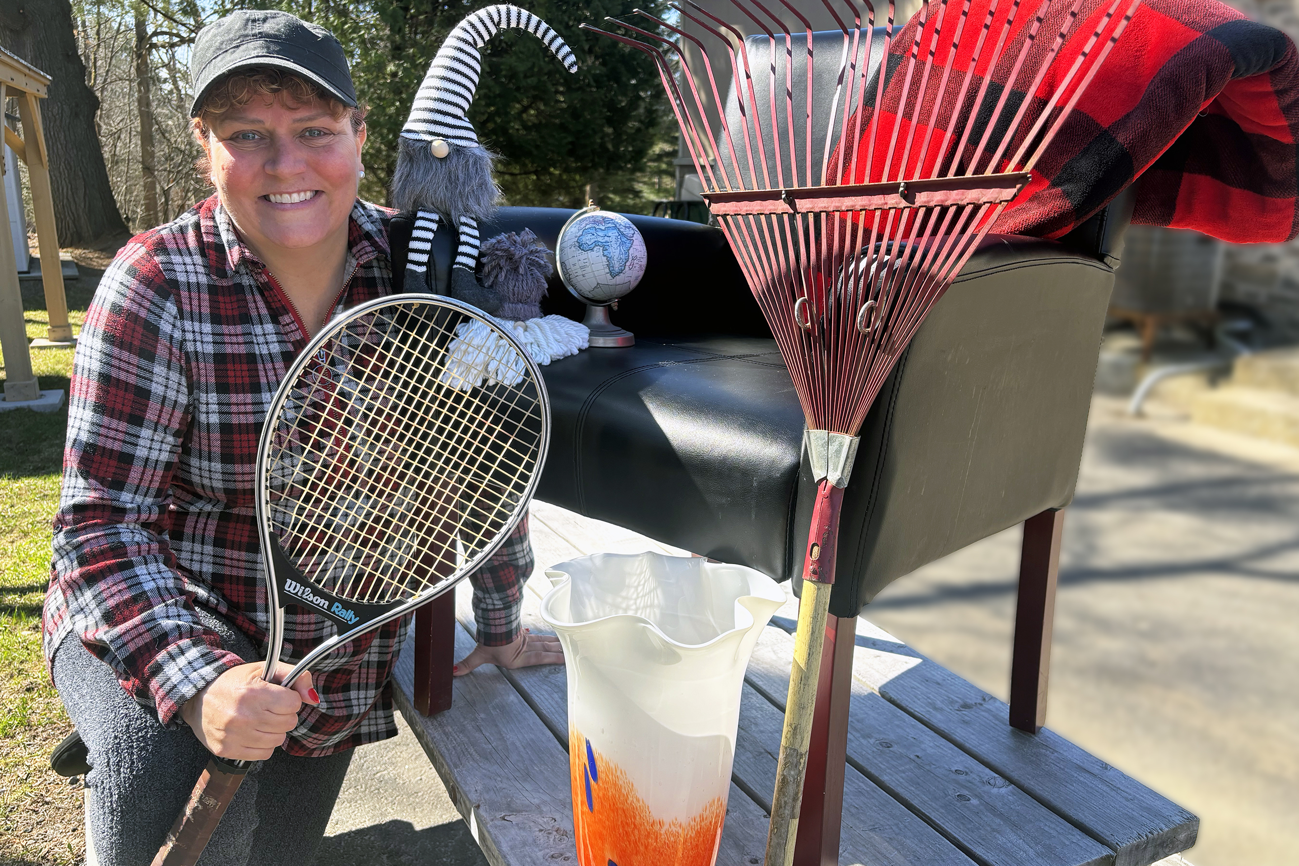 A woman in a ball cap and plaid shirt smiles while sitting outside on a picnic table with a chair, tennis racket, vase, rake and other home goods.