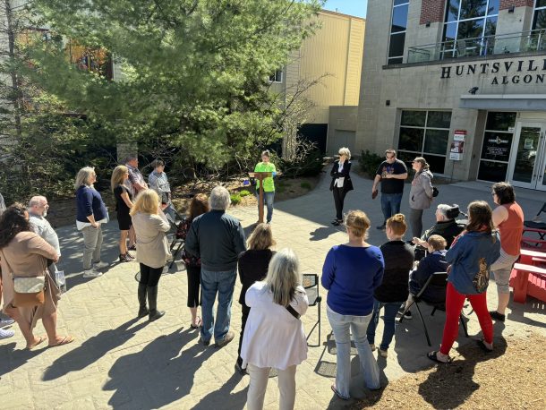 A crowd gathers around a woman who stands at a podium speaking