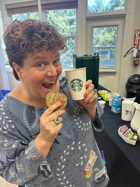 a woman, Jennifer, holds a cookie and a coffee near a table of refreshments.