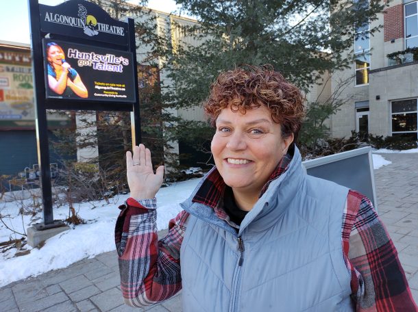 A smiling woman in a plaid shirt and blue outdoor vest stands in a snowy courtyard, Huntsville Civic Square, and gestures with her hand to a digital display screen that shows a singer and text that reads, "Huntsville's Got Talent."