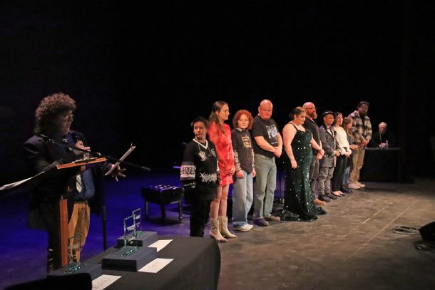 A man with a clipboard speaks into a microphone while a line of 10 people, the Top 10 Finalists, wait on stage.