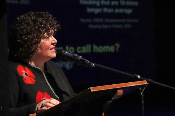 A woman with dark curly hair and a black sweater with large red poppies on it stands at a podium and speaks into a microphone.