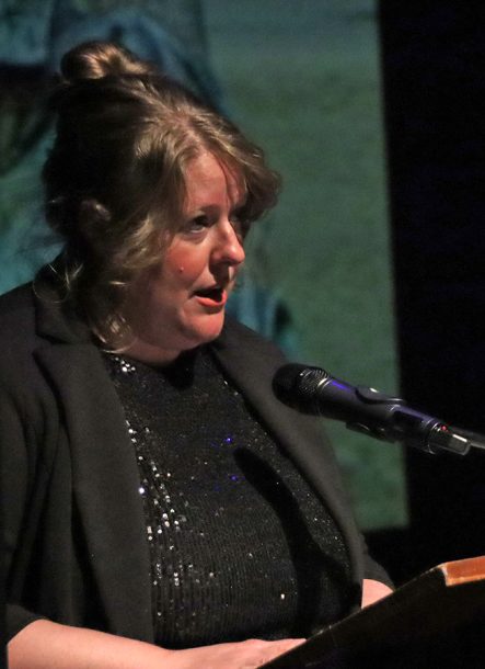 A woman with blonde hair, a sparkly black top and black cardigan stands at a podium and speaks into a microphone.