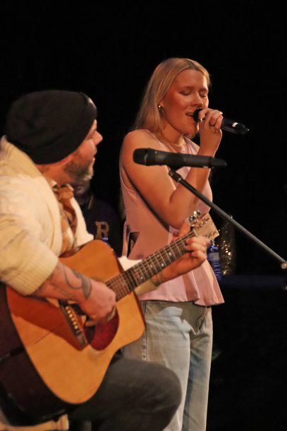 A man in a grey beanie and white sweater plays the guitar and sings into a microphone while a young woman in a pink top stands nearby singing into a microphone.