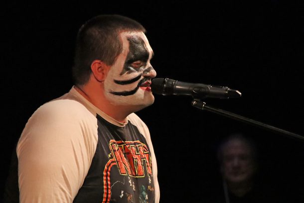 A man in a black and white T-shirt that features the glam-rock bank, Kiss, also wears black and white face paint like the band members while singing into a microphone