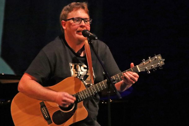 A man with red hair and glasses plays the guitar while singing into a microphone