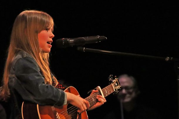 A young girl with a denim jacket and guitar sings into a microphone.