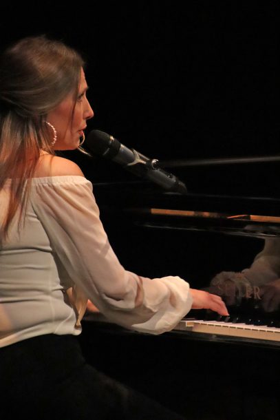 A woman in a white shirt plays a grand piano while singing into a microphone.