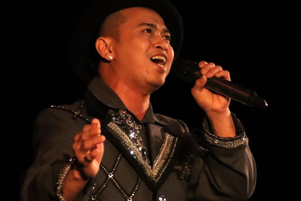 A man in a bowler hat and grey suit jacket decorated with rhinestones sings into a microphone