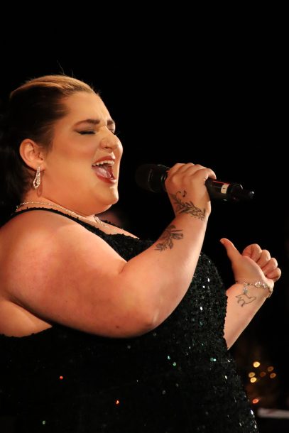 A woman in a green sequin ballgown leans back and sings into a microphone
