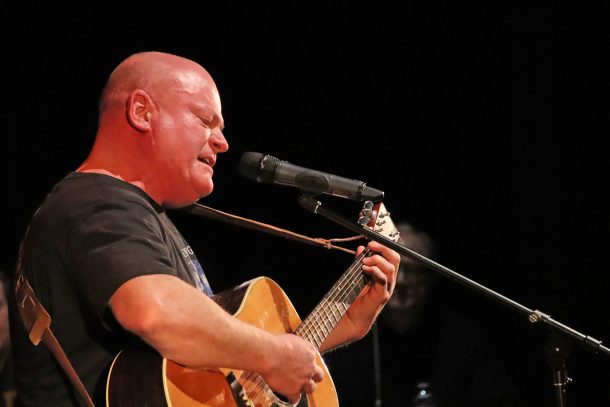 A man in a grey T-shirt and guitar sings into a microphone