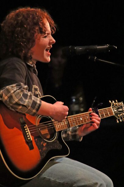A person with red curly hair, plaid shirt and grey T-shirt plays a guitar while singing into a microphone.