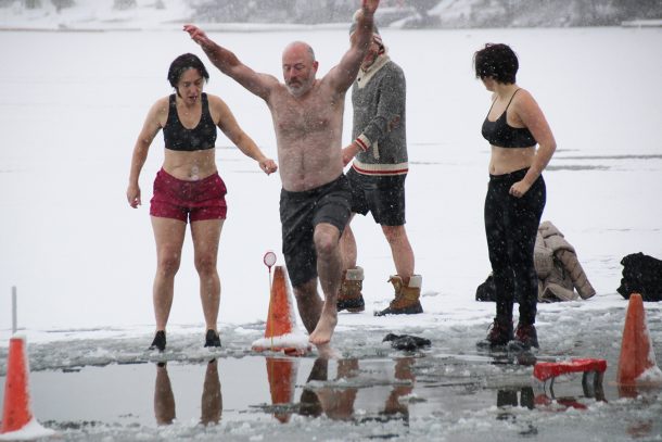 A man in swim trunks raises his arms as he leaps off a frozen lake into a purpose-built hole cut into the ice.