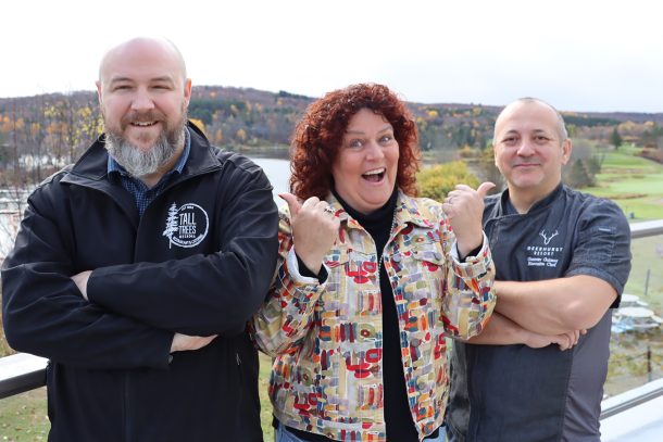 3 people, Randy, Jen, and Gus, stand outside on a patio with a lake and autumn trees behind them.