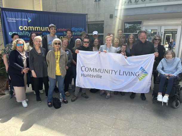 A large group of smiling people hold a white flag with the Community Living logo on it.