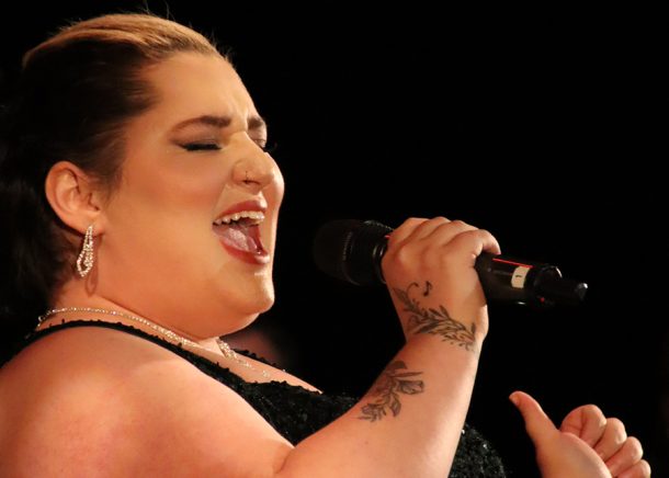 A woman in a green ballgown leans back and sings into a microphone she holds in her right hand.