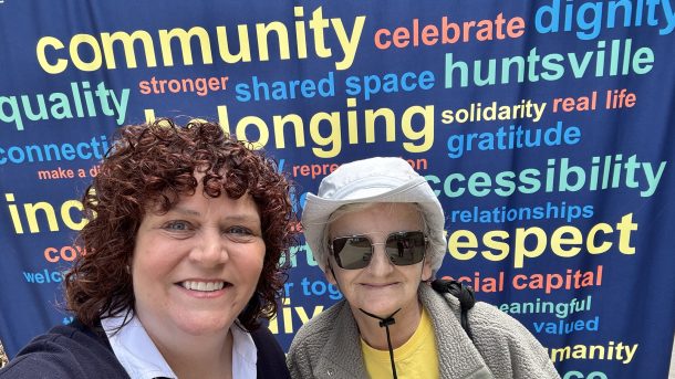2 smiling women, Jen and Cindy, pose for a selfie in front of a wordcloud banner filled with words like, "Community" and "Belonging."