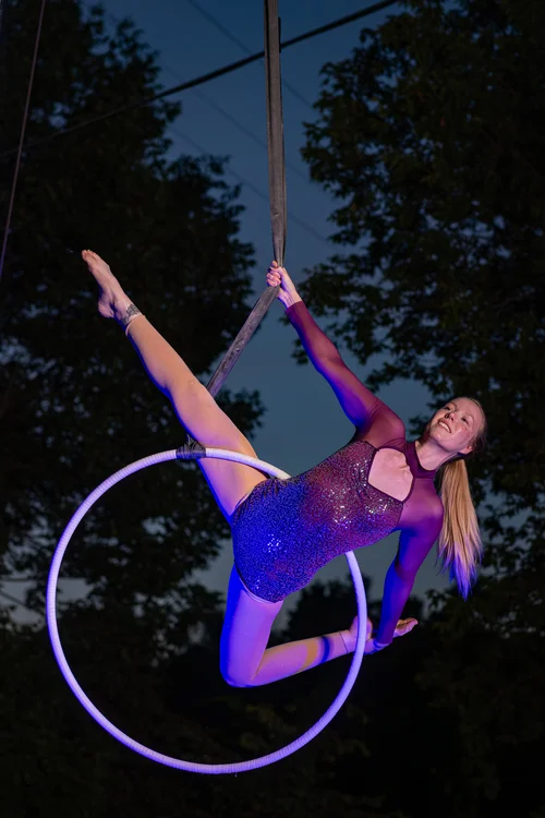 A smiling woman in a purple leotard lifts herself into the air using a suspended aerial arts hoop.