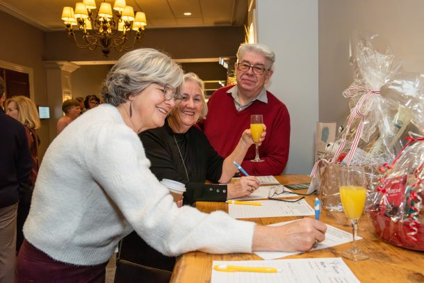 3 smiling people laugh as 1 woman signs a silent auction bid sheet in front of a gift basket