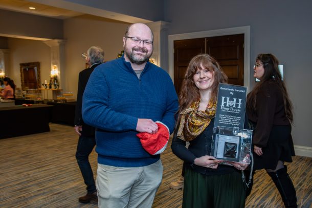 a man and a woman smile as they hold a small display case that reads The Hunt House