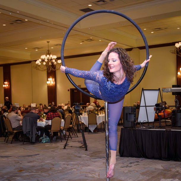 An aerial artists in a blue leotard lifts her leg behind her as she leans forward into a lollypop hoop.