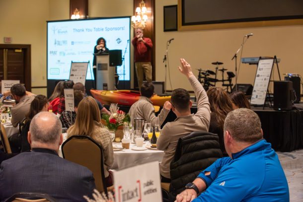 A man raises his hand in a crowd as an auctioneer on stage points in his direction