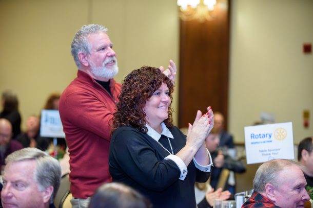 A man and a woman smile as they stand in a crowded room while applauding