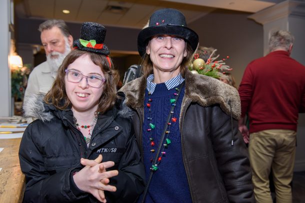 2 women, a mother and a daughter, smiling and wearing festive hats and necklaces.