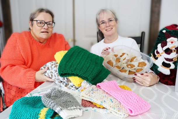 2 smiling women seated at a dining room table. 1 holds knitted scarves and the other holds a container of homemade muffins.