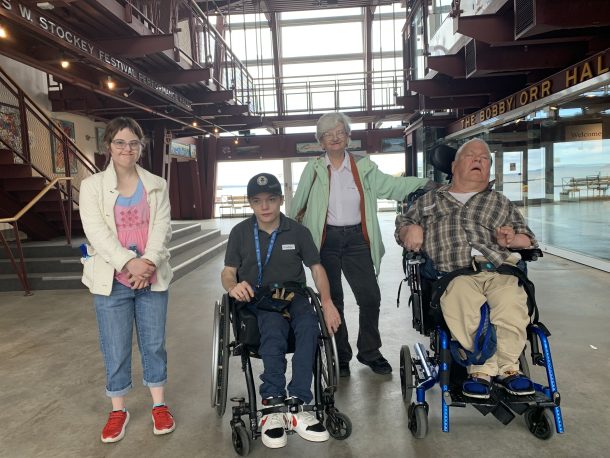 4 people, 2 standing and 2 sat in wheelchairs, pose for a group photo in a building with floor to ceiling windows behind them.