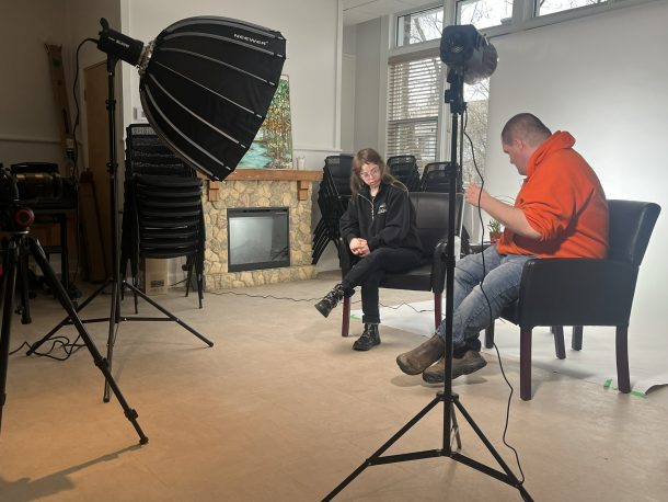 A woman and a man seated in front of a white backdrop with film lighting shining on them.