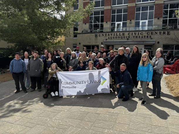 A large group of people of all ages and abilities stand outside the Huntsville Civic Centre of a sunny day, holding a large white flag decorated with the Community Living logo.