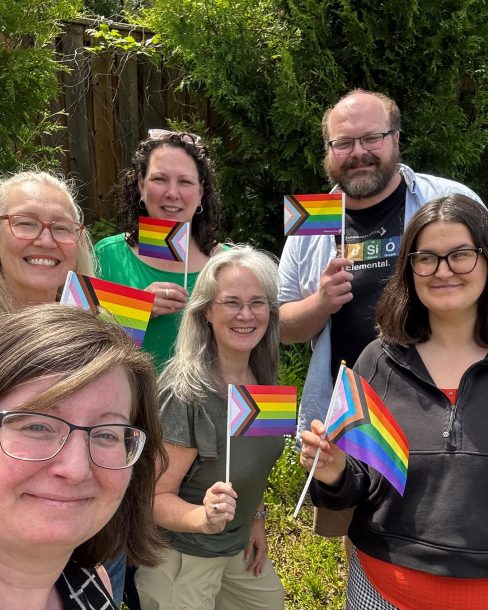6 smiling adults outside on a sunny day holding paper progress pride flags.