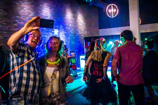 A man and a woman wearing glowing headphones and neon glow sticks take a selfie in a darkened room while other people wearing glowing headphones dance nearby.