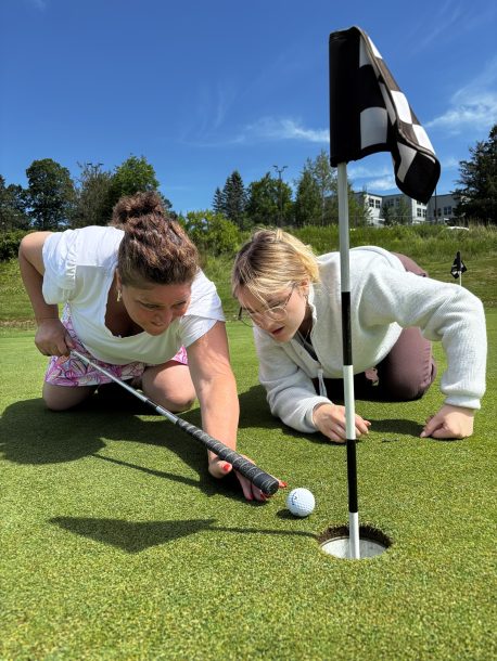 2 women kneel on a golf green and peer into the pin. 1 women holds a putter like a pool cue and readies to poke a golf ball into the golf green cup.