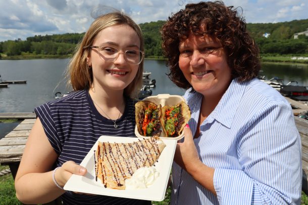 2 women at the waterfront. 1 holds 2 tacos, while 1 holds a plate full of crepe.