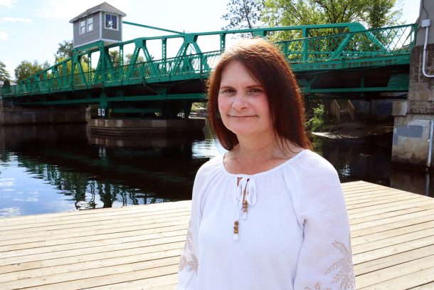 A woman, Sherri-Lynn, stands on a dock near a green swing bridge in downtown Huntsville on a sunny day.