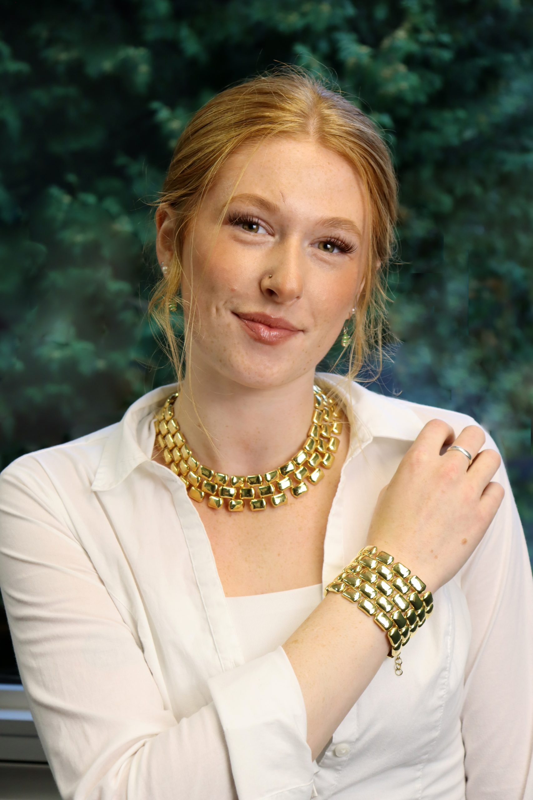 A woman with red hair and a white shirt poses while wearing a gold necklace and matching bracelet.