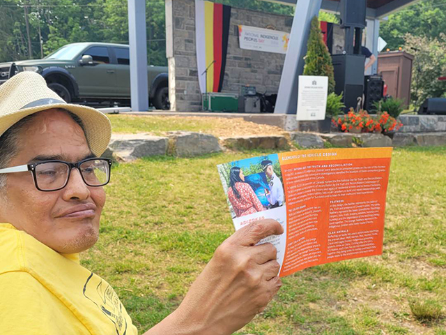 A man seated in a public park near a stage decorated for National Indigenous People's Day.