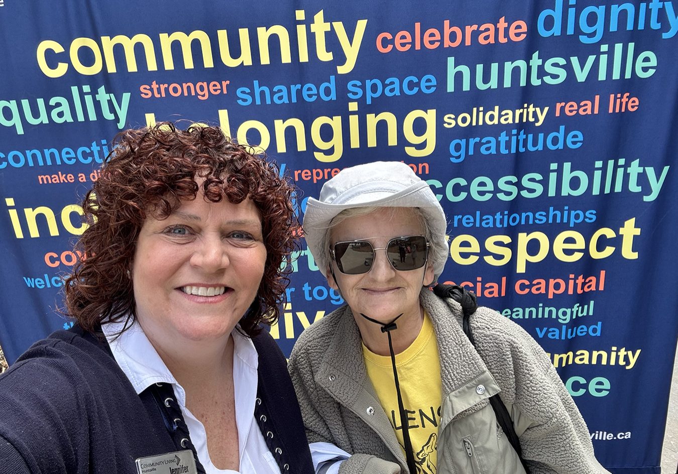 2 women smile as they pose for a selfie in front of a dark blue banner that is covered in words like "Community" and "Inclusion."
