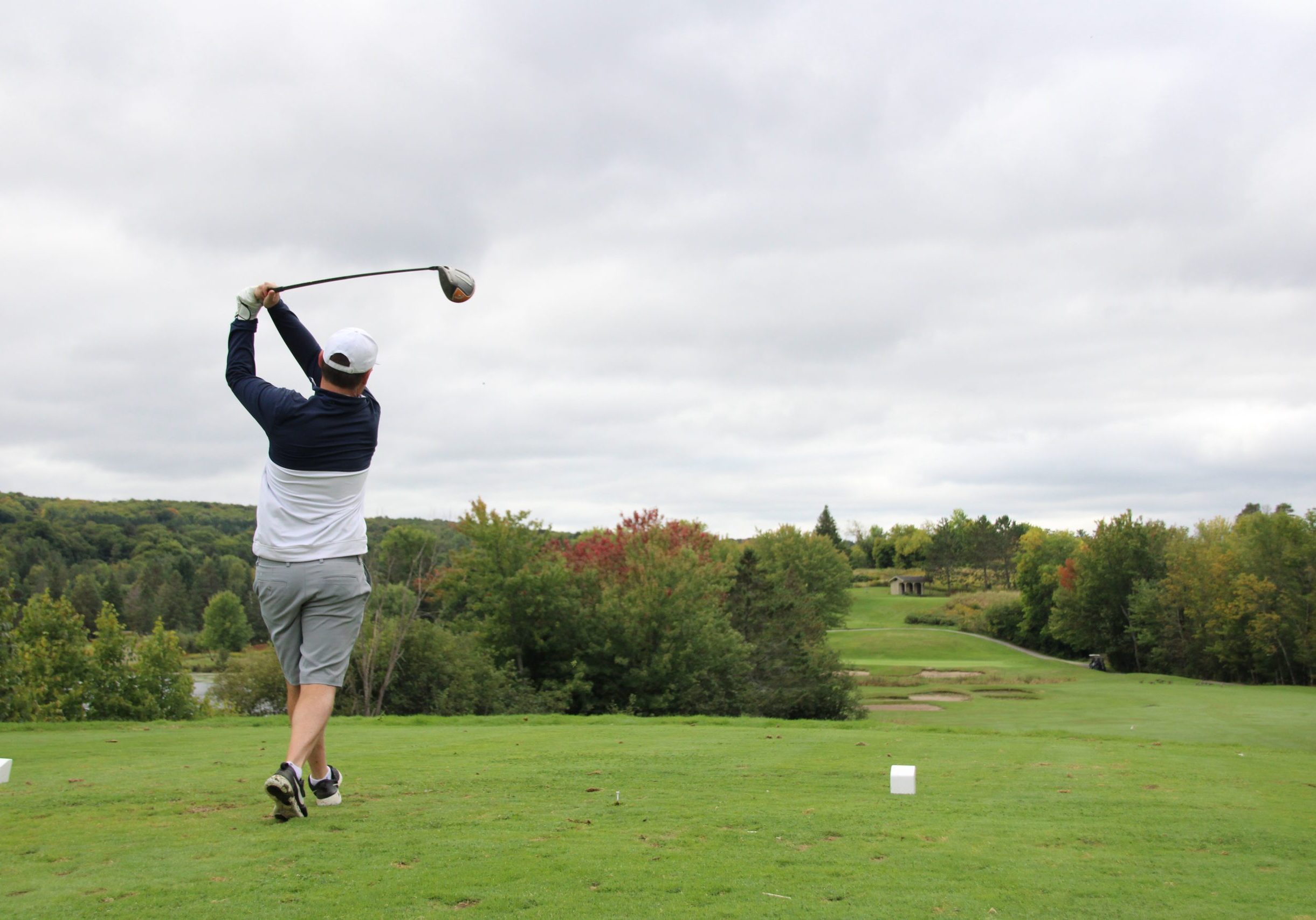 A man swings through a tee off while on a green golf course surrounded by trees.